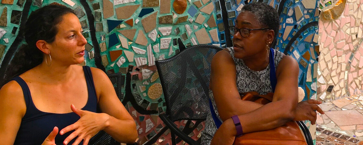 Two women having a discussion outside in front of a tile mosaic wall